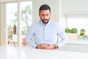 A man in a button-up shirt is closing his eyes and holding his stomach with both hands.