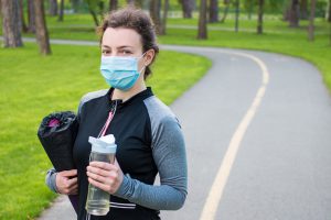 A woman in athletic apparel stands on a track wearing a face mask and holding a water bottle.